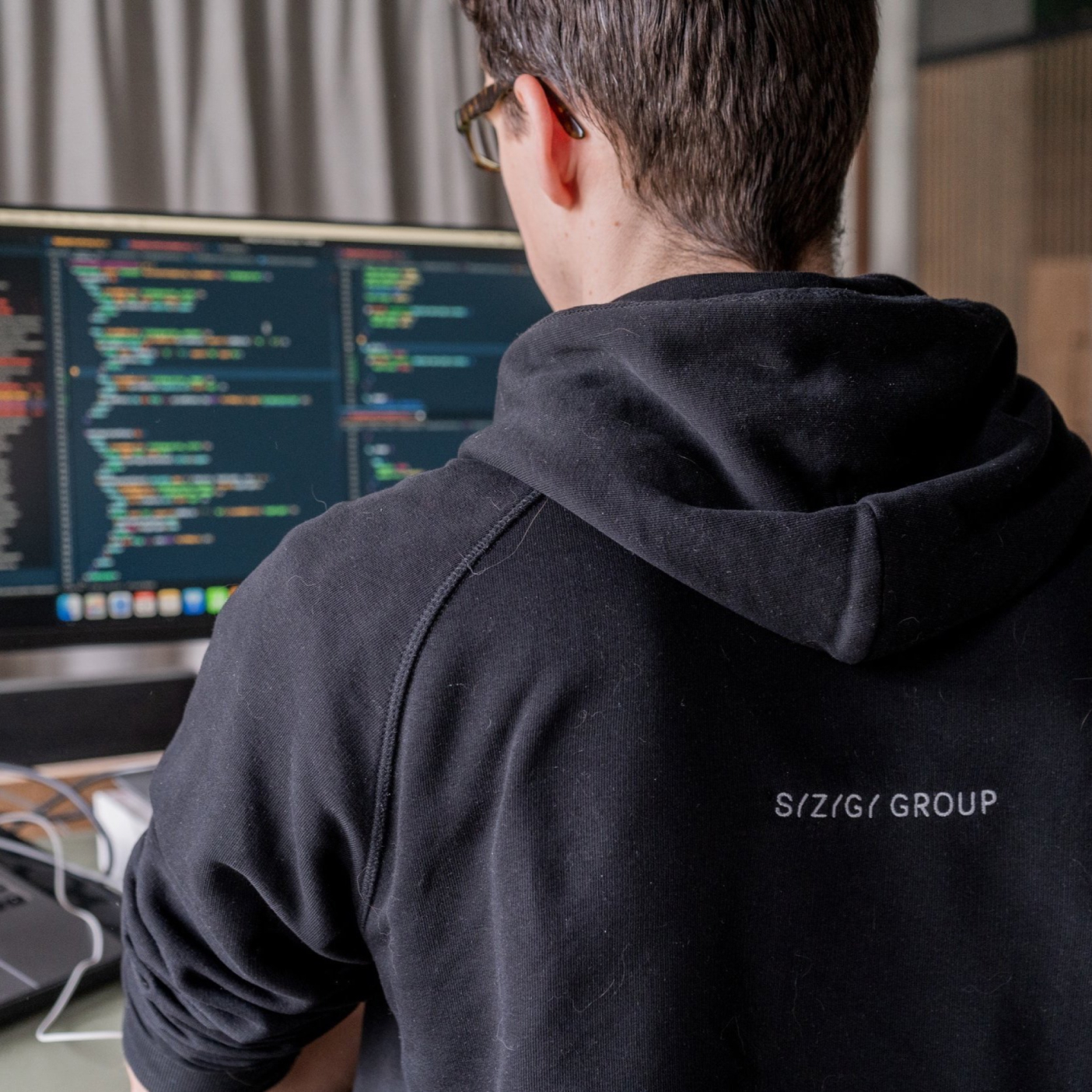 A man in a black hoodie working at the computer. Lines of code are visible on the computer screen.